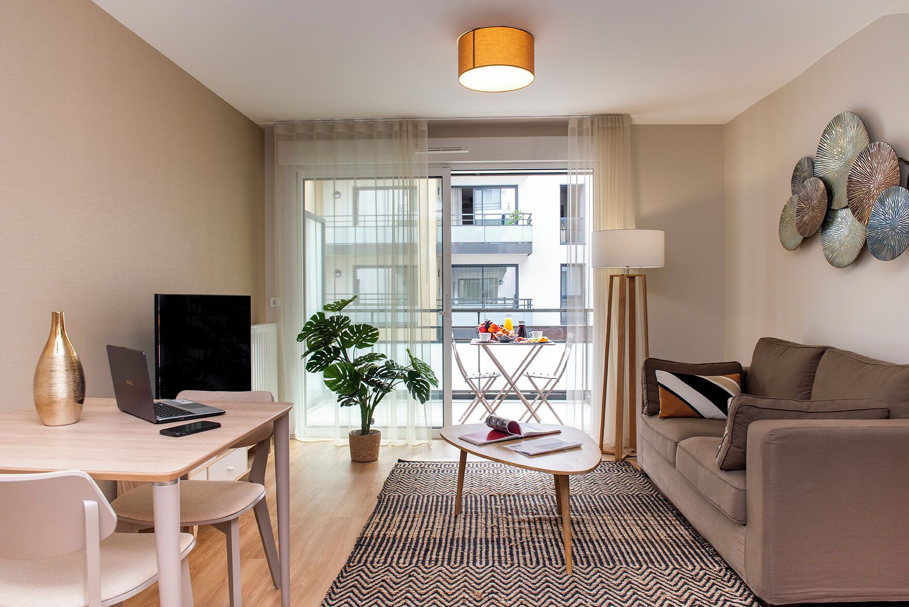A modern living room with a sofa, a dining table, a laptop, a plant, and a view of the outside balcony.