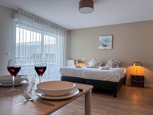 A hotel room with a bed, bedside lamps, a dining table with wine glasses and a bowl, and a window with blinds.