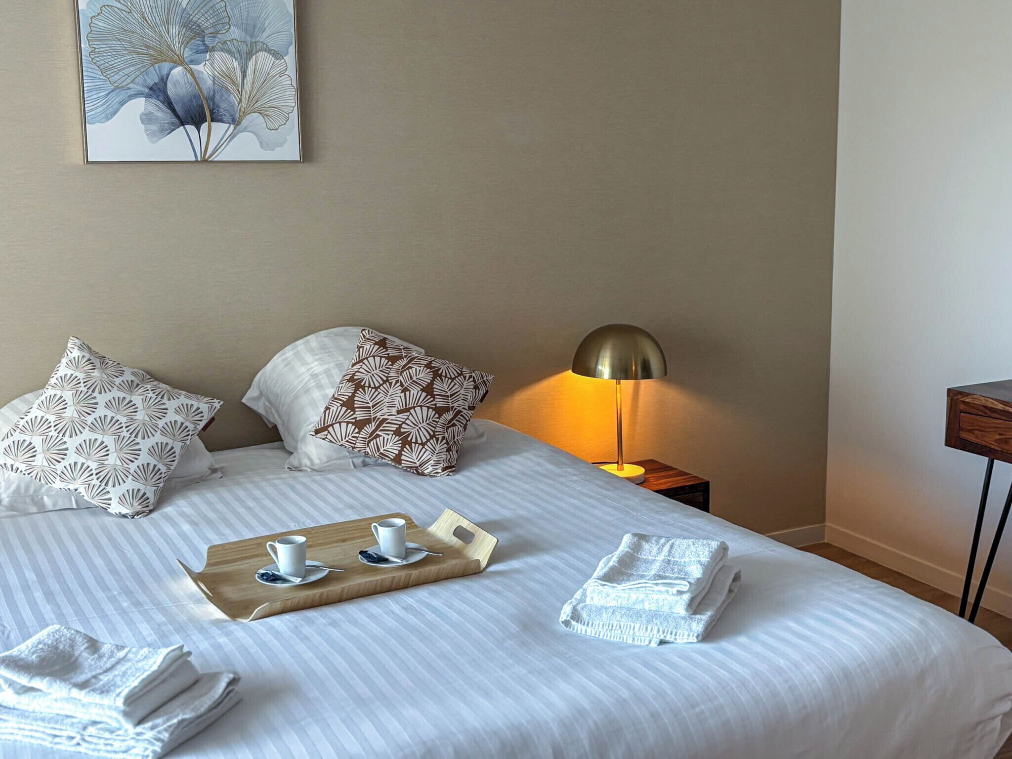 Een keurig opgemaakt bed met kussens, een nachtkastje met een lamp, een dienblad met bekers en een houten plank, en opgevouwen handdoeken.
