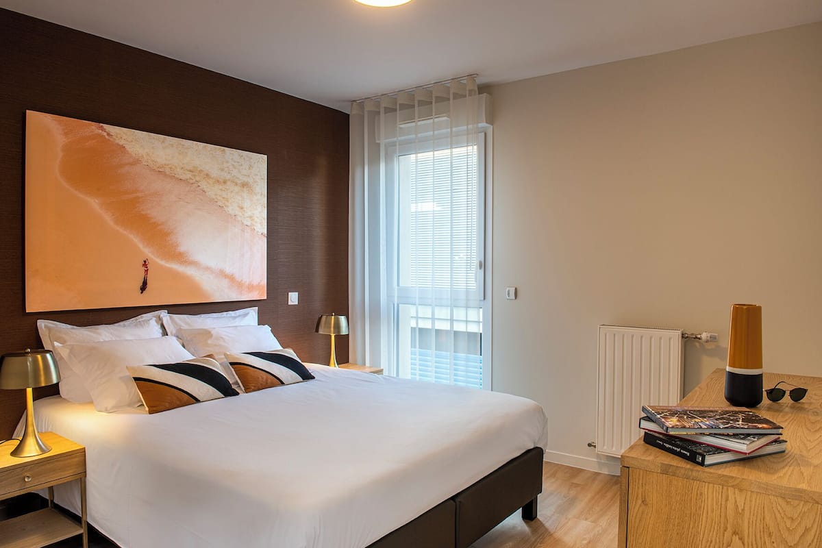 Una camera d'albergo moderna con un letto grande, un comodino con una lampada, un tavolino in legno con un libro e occhiali da sole, e un grande quadro astratto appeso al muro.