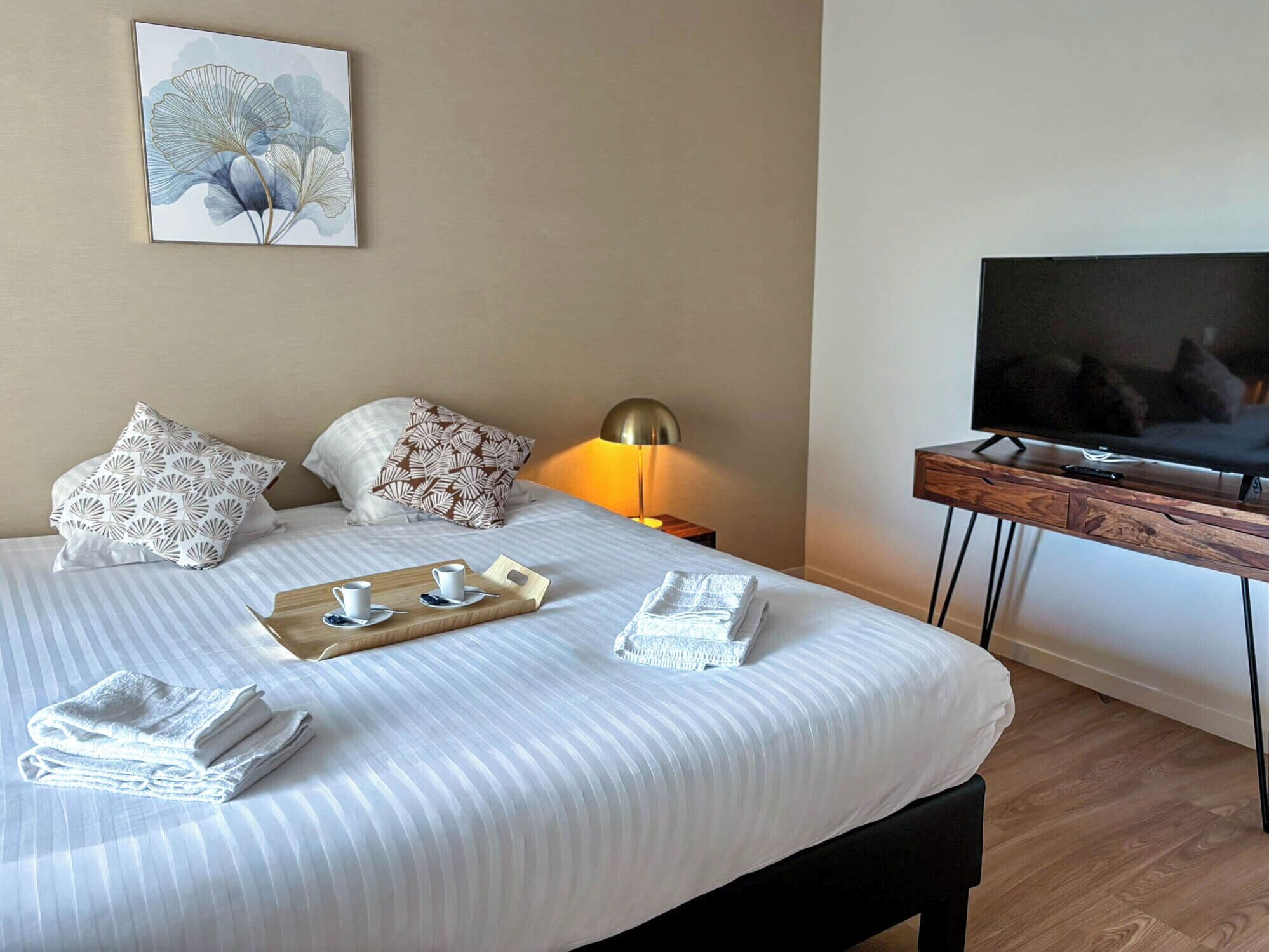A neatly made bed with pillows, a tray with cups and a remote, a TV on a wooden stand, and a painting on the wall.