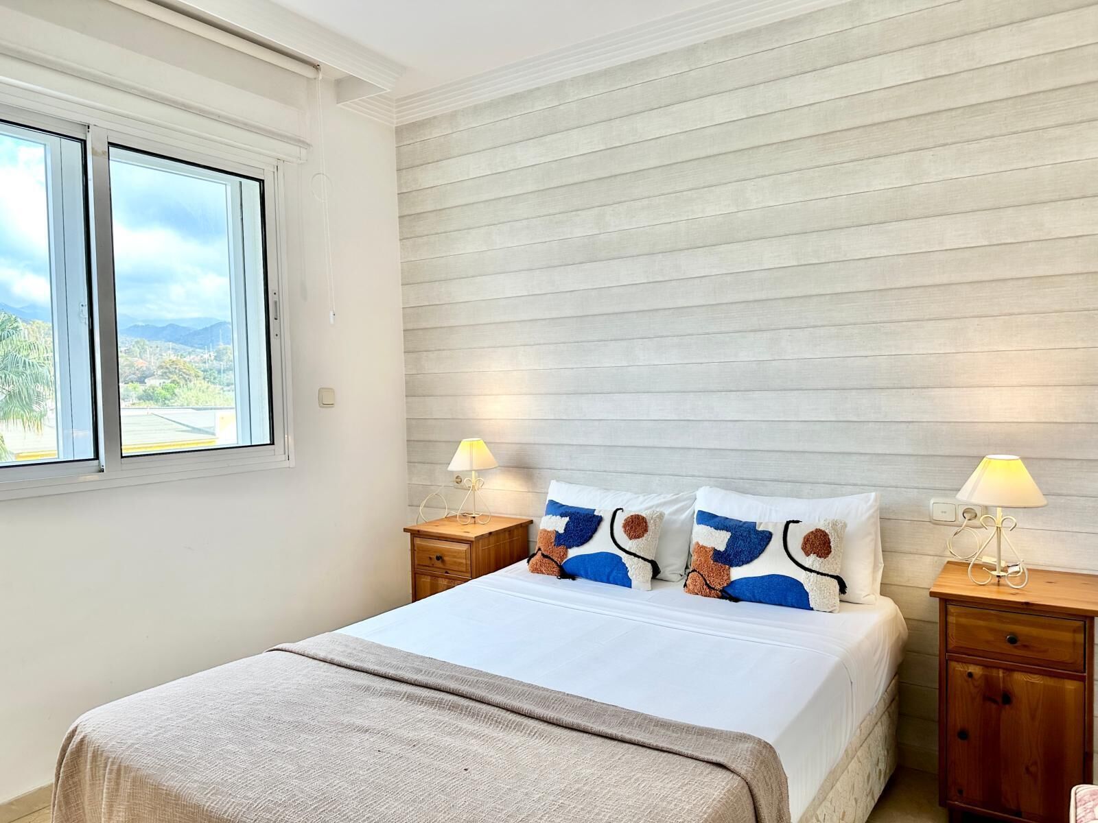 A bedroom with a bed, two bedside tables with lamps, a window with a view of greenery, and a wooden headboard.