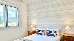 A bedroom with a bed, two bedside tables with lamps, a window with a view of greenery, and a wooden headboard.