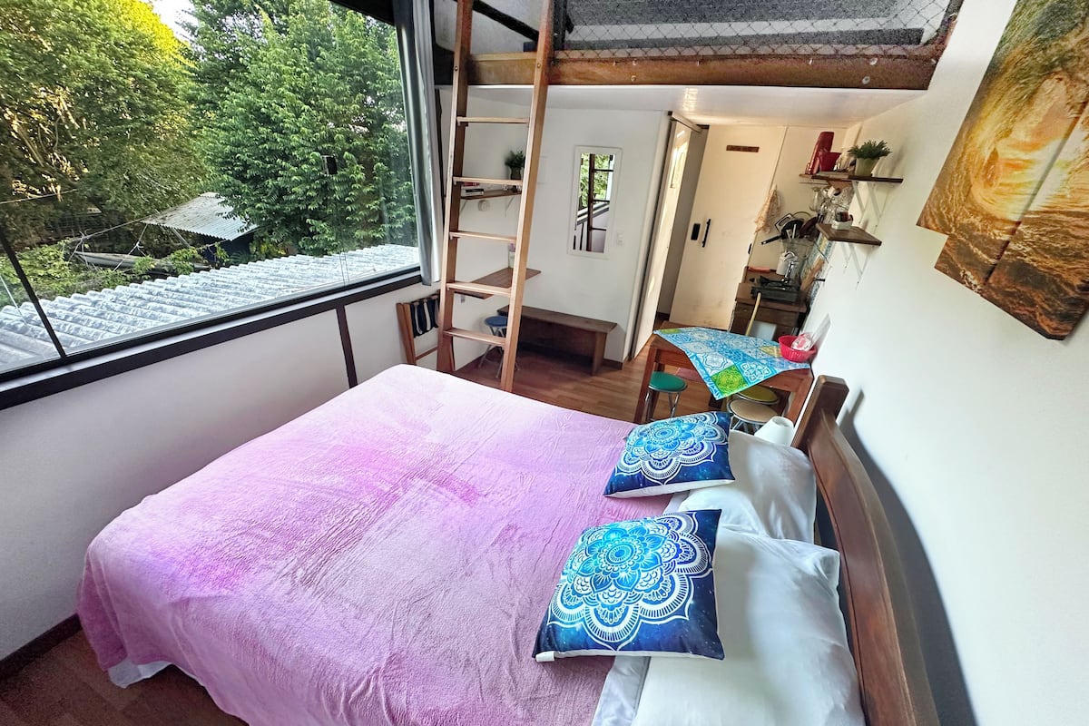 Ein Schlafzimmer mit Bett, einer Leiter, einem Schreibtisch und Blick auf Bäume.