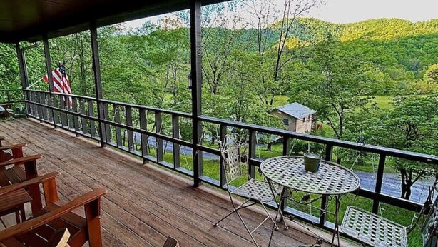 A covered outdoor deck with wooden chairs and a metal table, overlooking a lush green landscape with hills and a small building in the distance.