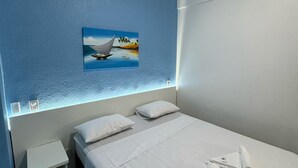 A modern hotel room with a large bed, white linens, a painting of a boat on water, a nightstand, and a small shelf. - Hotel Américas (Fortaleza)