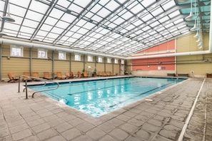 Una piscina interior con techo de vidrio, sillas de descanso y suelo de baldosas.