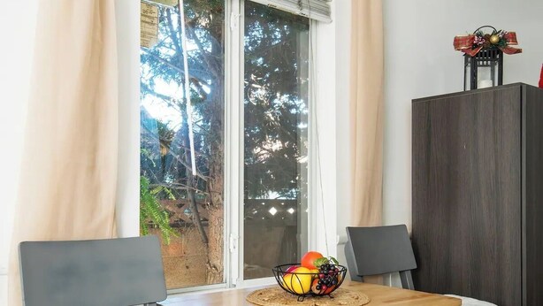 Un petit coin repas comprenant une table, deux chaises, un panier de fruits, un meuble et une fenêtre avec des rideaux.