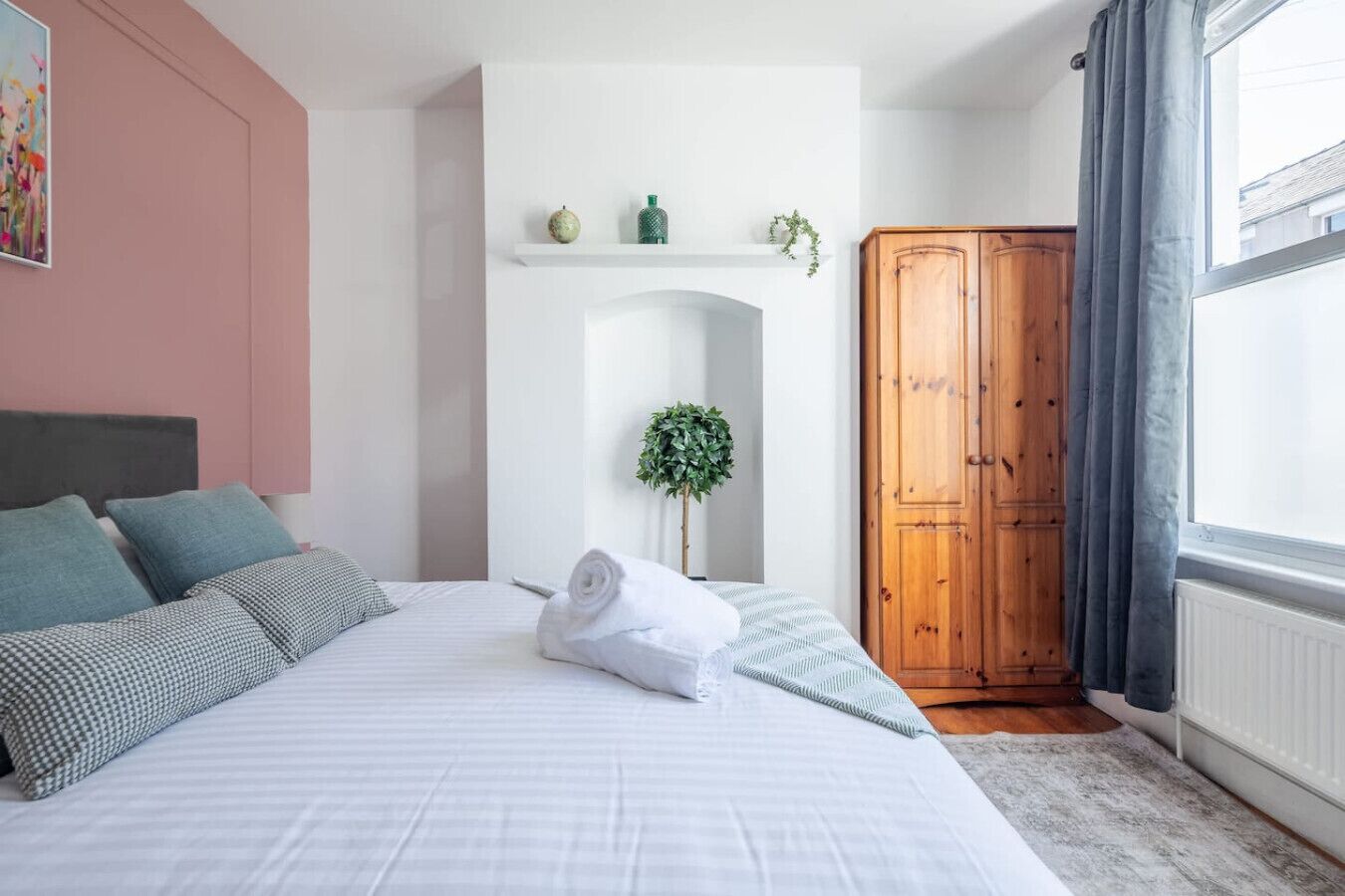 Ein Schlafzimmer mit einem Bett, Kissen, einem gerollten Handtuch, einem Holzschrank, einer Pflanze und einem Fenster mit Vorhängen.