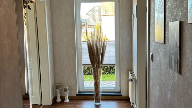 Ein Flur mit einer hohen Vase voller Trockenpflanzen, einer zur Außenseite führenden Tür und einem Heizkörper an der Wand.