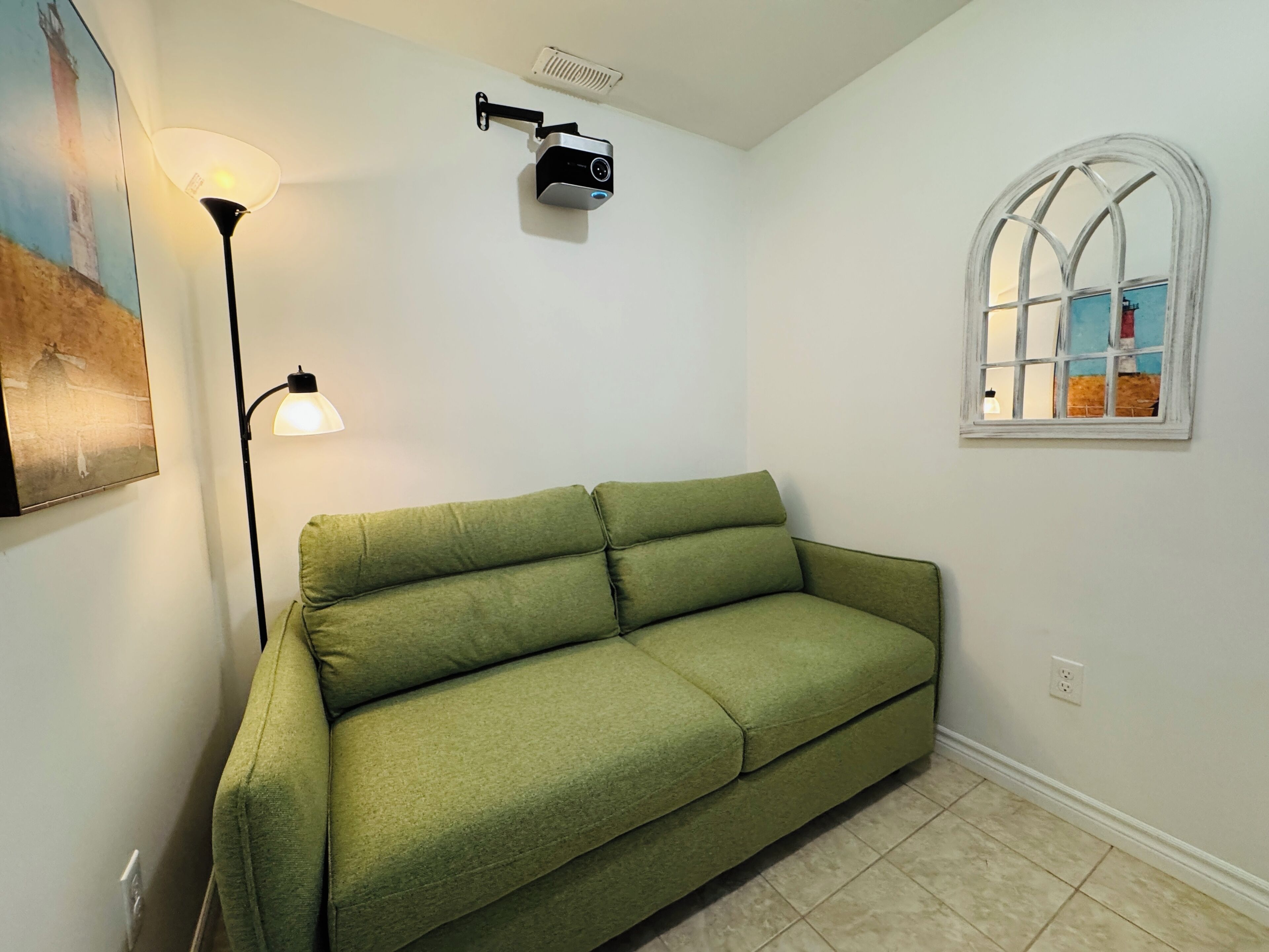A green sofa, a floor lamp, a projector, a window with a view, and a painting on the wall.