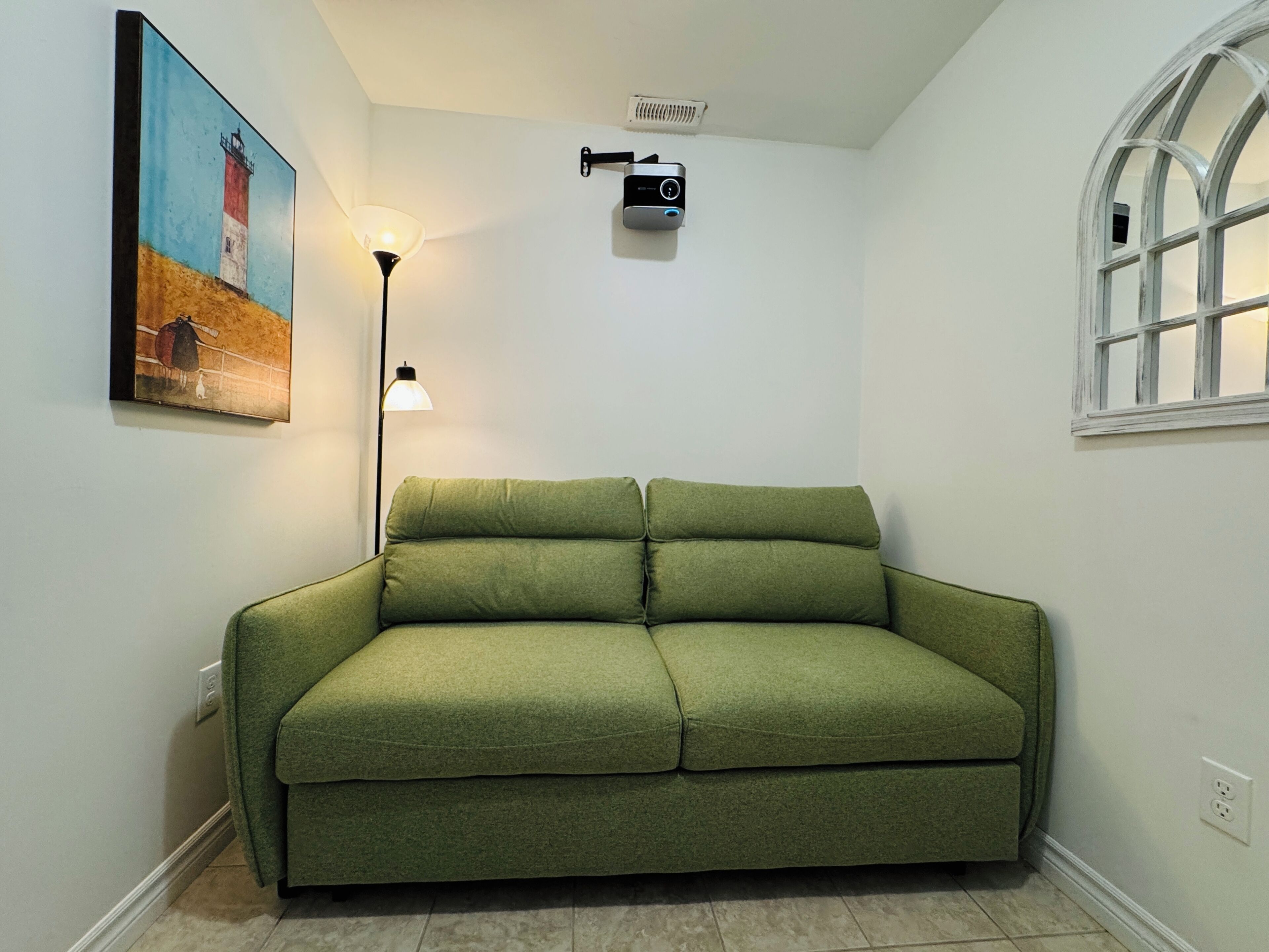 Une pièce avec un canapé vert, un tableau représentant un phare, une lampe, un projecteur et une fenêtre.