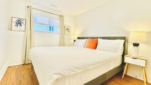 A neatly made bed with white linens and a few pillows, a bedside table with a lamp, and a window with curtains. - Lovely King Basement Suite & Small Cinema & Fireplace (Halifax)