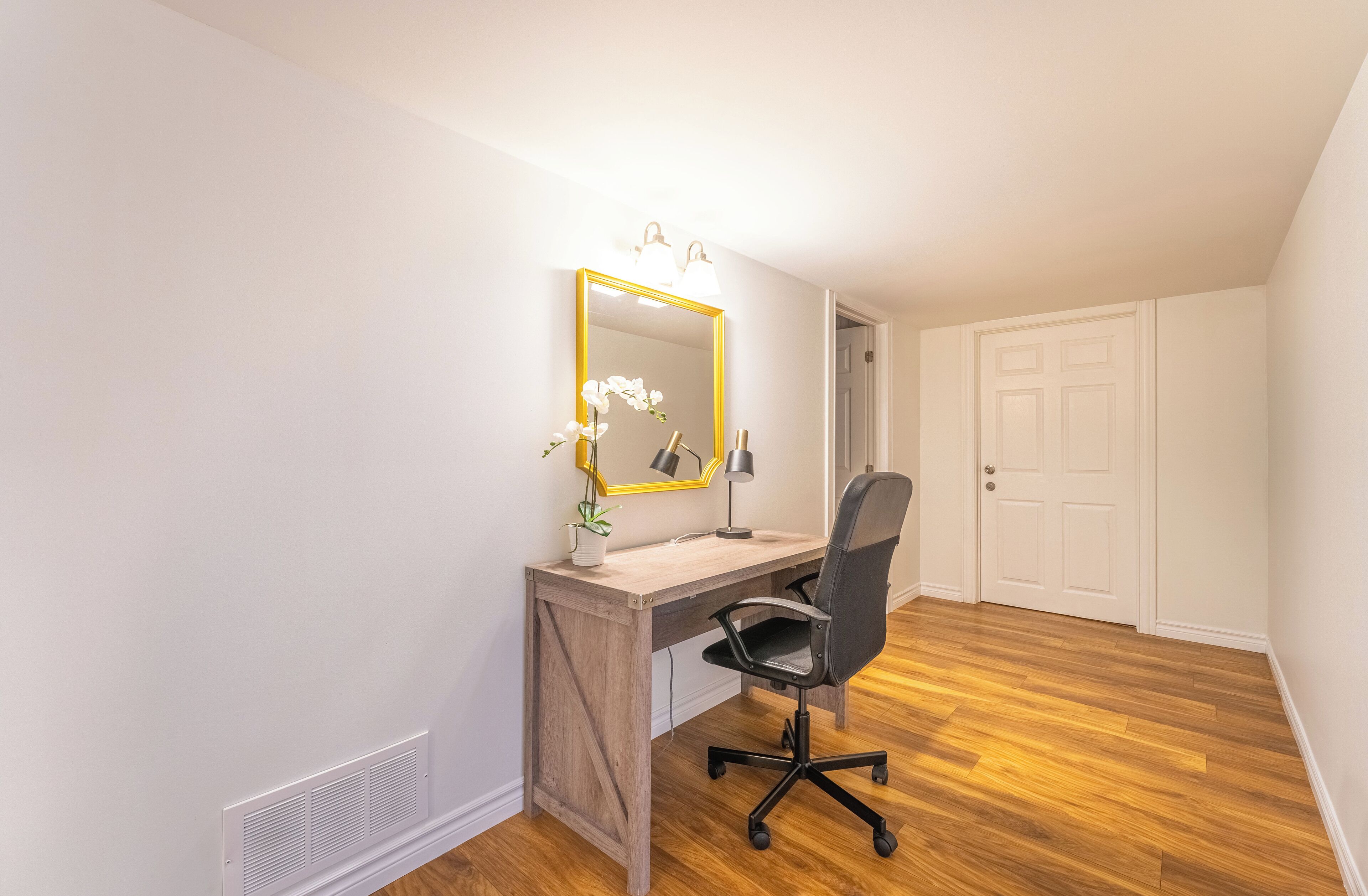 Une pièce avec un bureau en bois, une chaise de bureau noire, un miroir avec un cadre doré et une petite plante en pot.
