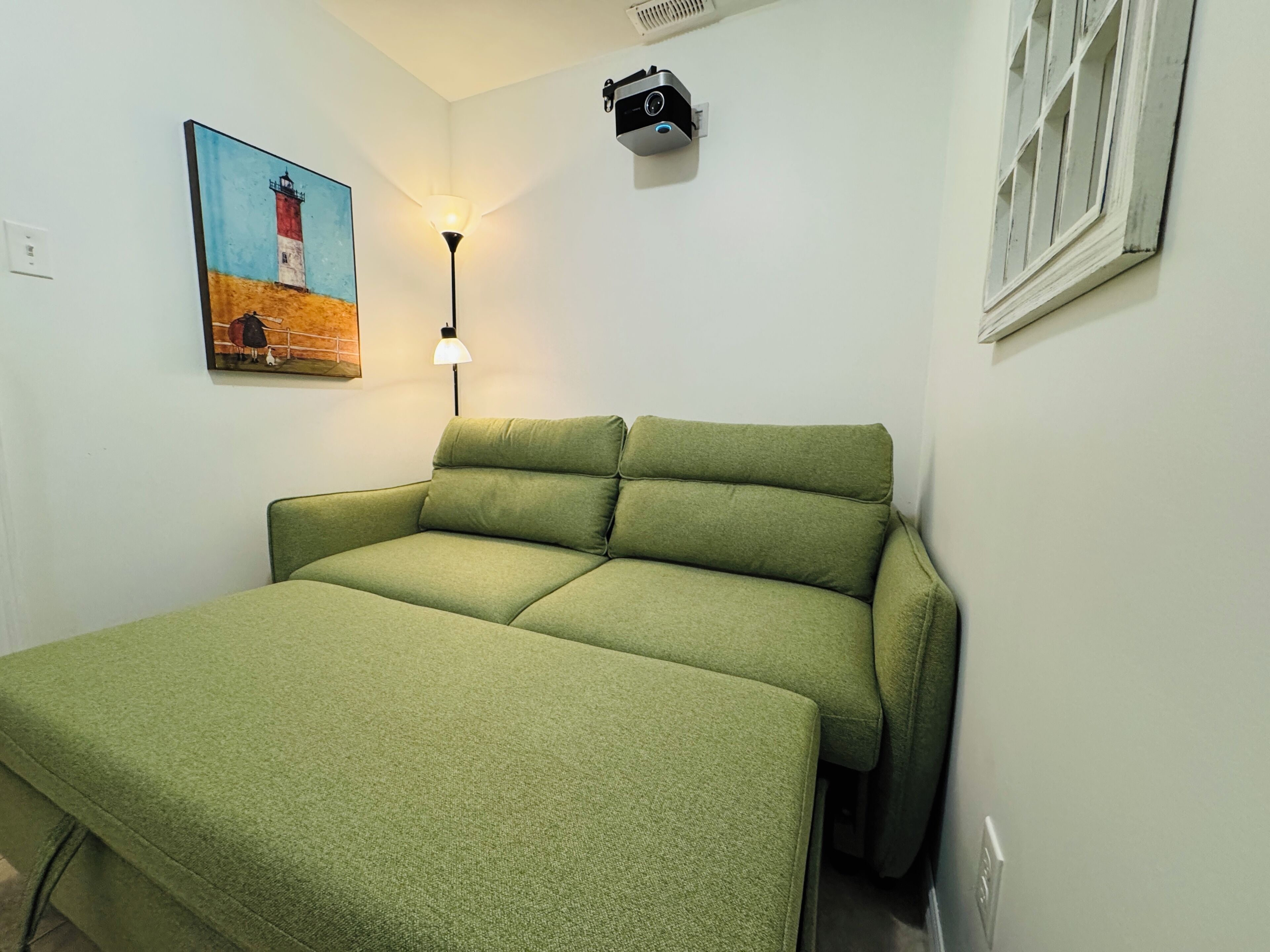 Una stanza con un divano letto verde, un quadro raffigurante un faro, una lampada da scrivania e un proiettore montato a parete.