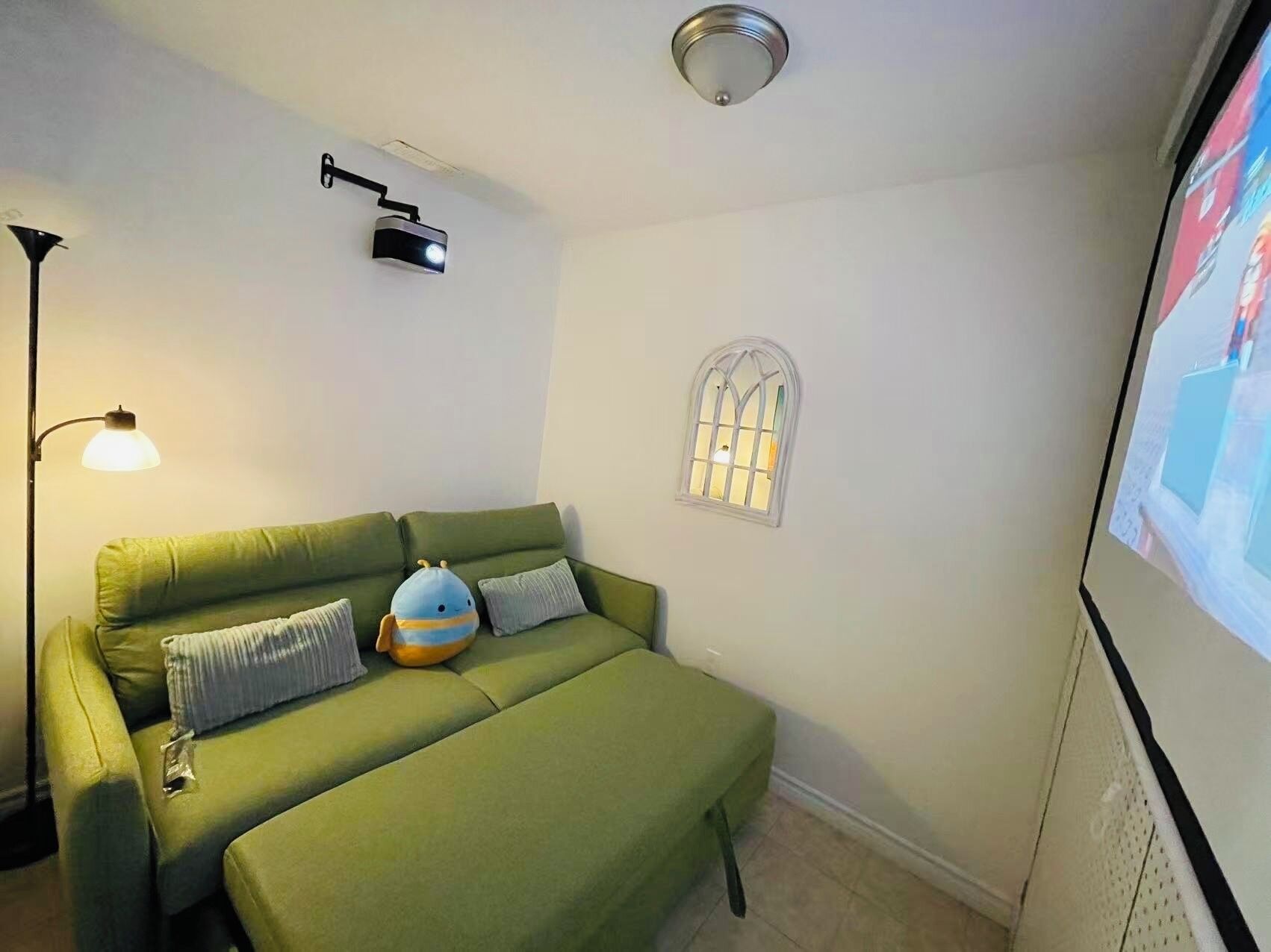 Un canapé vert avec un petit jouet en peluche bleu, un projecteur fixé au mur, une lampe sur pied et une fenêtre ornée d’une grille décorative.
