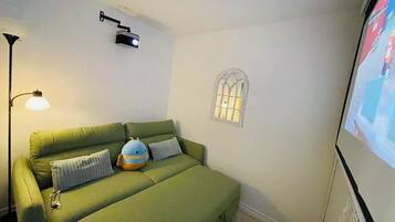 Un canapé vert avec un petit jouet en peluche bleu, un projecteur fixé au mur, une lampe sur pied et une fenêtre ornée d’une grille décorative.