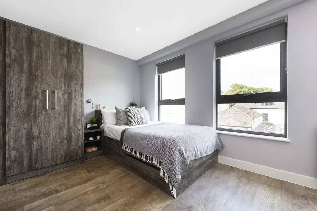 Une chambre à coucher comprenant un lit, une armoire, une table de chevet et une fenêtre donnant sur des immeubles.