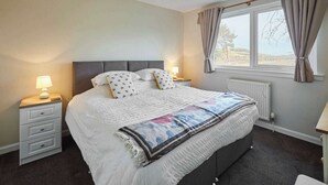A bedroom with a large bed, two bedside tables with lamps, a window with curtains, and a radiator. - Host & Stay | Audrey's Cottage (Kirk Yetholm)