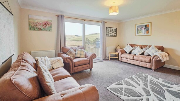 A spacious living room with two leather sofas, a patterned rug, and a view of the countryside through a large window. - Host & Stay | Audrey's Cottage (Kirk Yetholm)