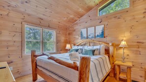 Un dormitorio de cabaña de madera con una cama, dos mesitas de noche, un ventanal y un cuadro en la pared.