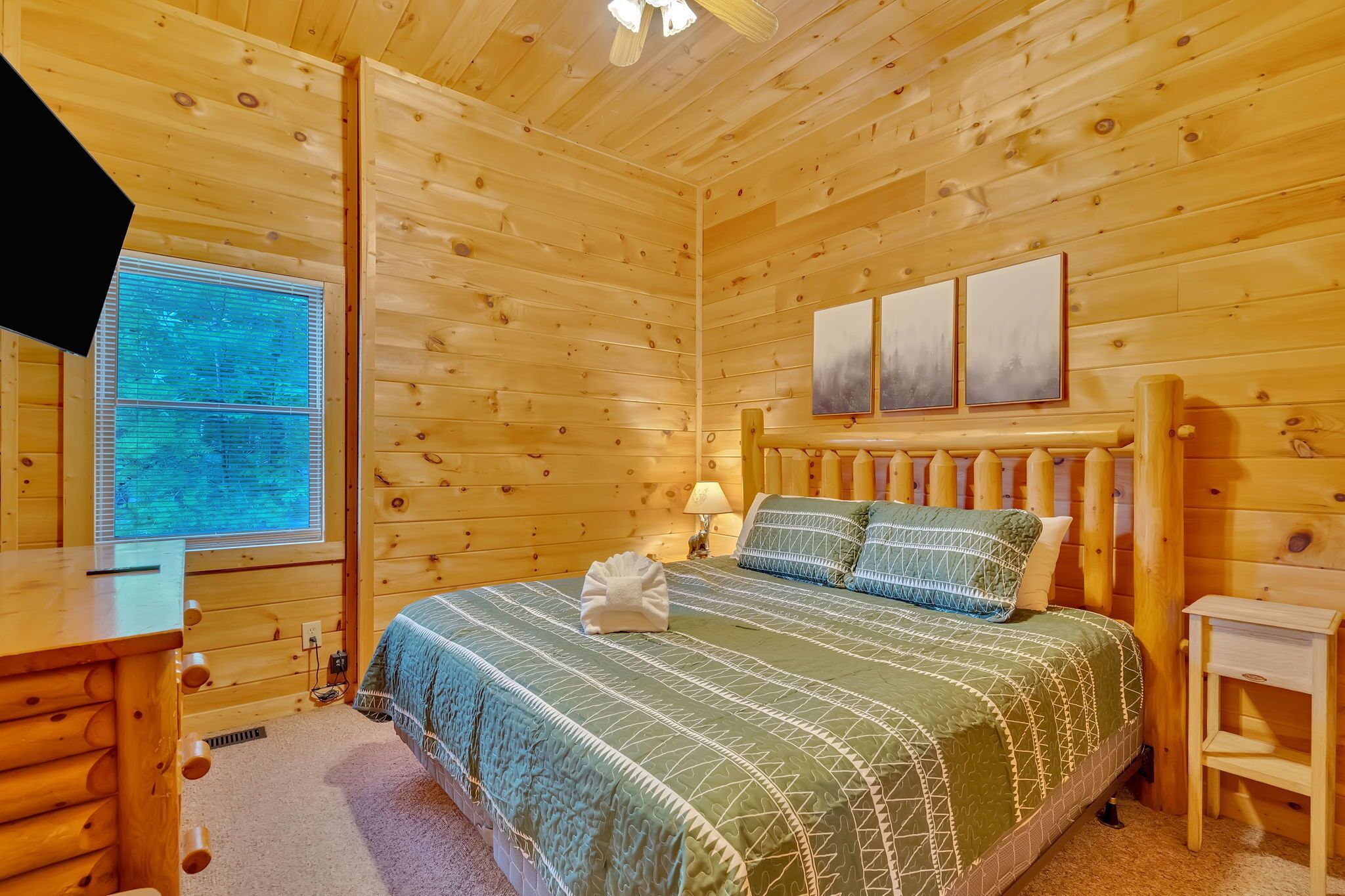 Un dormitorio de cabaña de madera con una cama, una mesita de noche, un ventanal y un televisor montado en la pared.