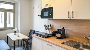 A compact kitchen with a microwave, toaster, and coffee maker on the counter, a sink, and a dining area with a table and chair. - Central apartment for 4 people with kitchen, WiFi & private parking space (Meißen)