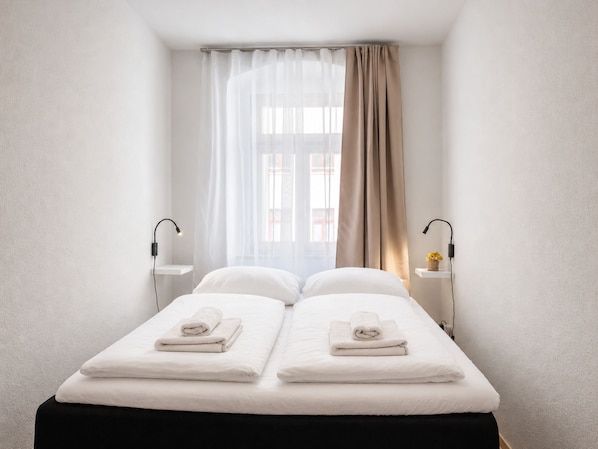 A hotel room with a bed, two bedside tables, a window with sheer curtains, and a lamp on each table.