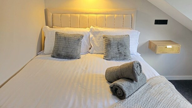 A neatly made bed with white linens and two gray pillows, a small bedside table with a radio, and a sloped ceiling with a single light fixture. - Warm & Cosy Home in Glasgow (Glasgow)