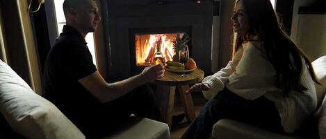 A couple seated by a fireplace with a table in front of them, holding drinks.