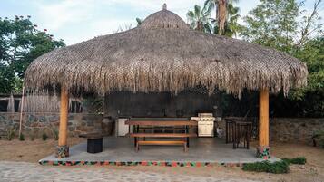 A thatched-roof structure with a stone wall, a wooden bench, and a small table, surrounded by a stone-paved area.