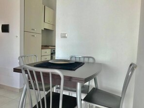 Een eethoek met een ronde tafel en vier stoelen, een keuken op de achtergrond en een lichtschakelaar aan de muur.