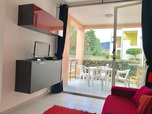 A room with a TV on a cabinet, a red sofa, a balcony with a table and chairs, and a view of greenery outside.
