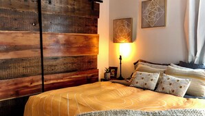 Een slaapkamer met een houten hoofdbord, een bed met gestreept beddengoed, een bedlampje en twee geometrische kunstwerken aan de muur.