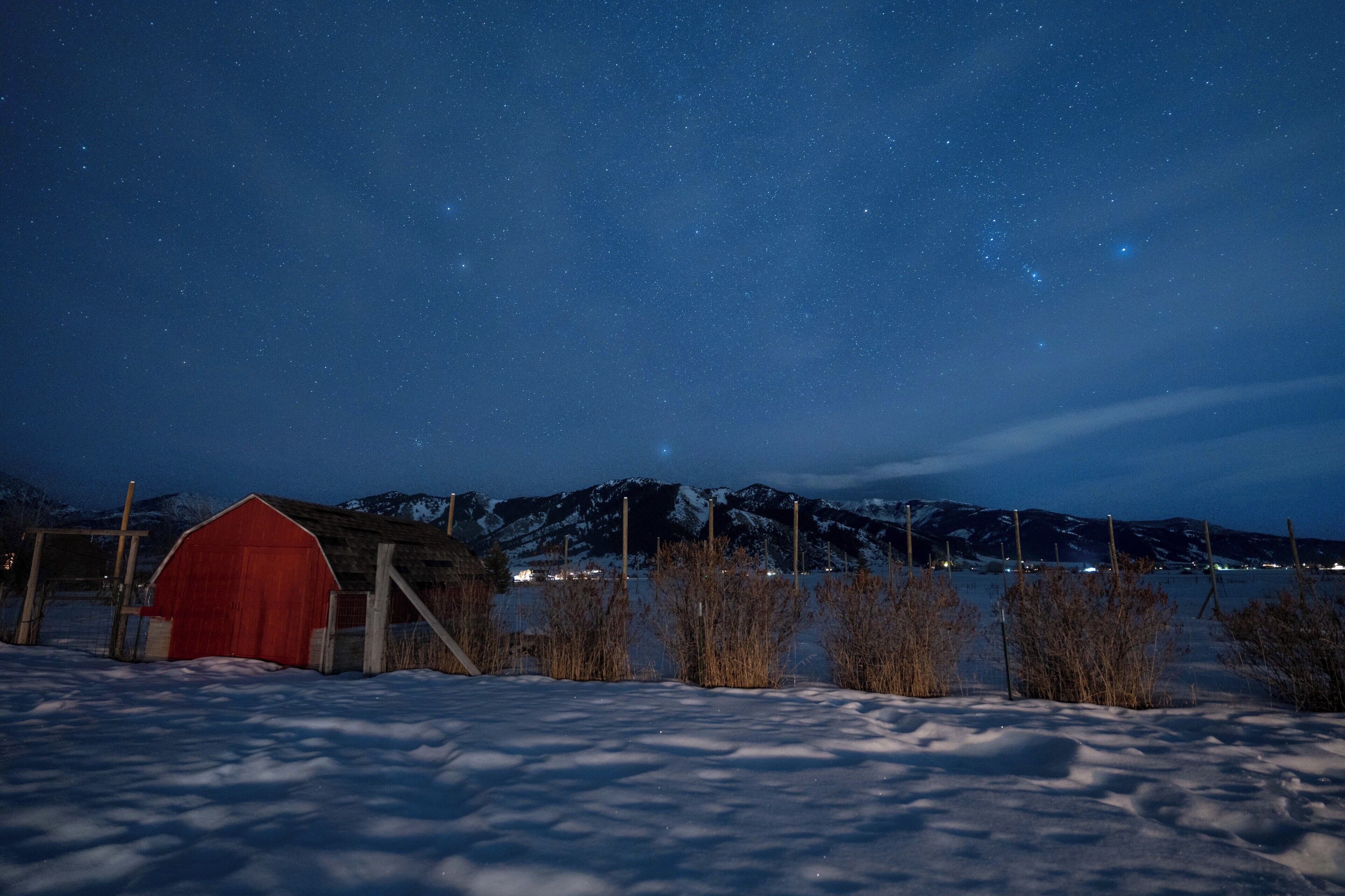 Ein rotes Bauerngebäude in einer verschneiten Landschaft mit sternenklarer Nacht und Bergen im Hintergrund.