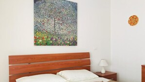 Une chambre à coucher avec une tête de lit en bois, un lit recouvert d’une literie blanche et beige, une table de chevet avec une lampe, et un tableau représentant un arbre et des fleurs accroché au mur.