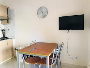 A dining area with a wooden table and chairs, a clock on the wall, and a flat-screen TV mounted above the table. - Pizzo Beach Club One Bedroom Apartment 128G (PIZZO)