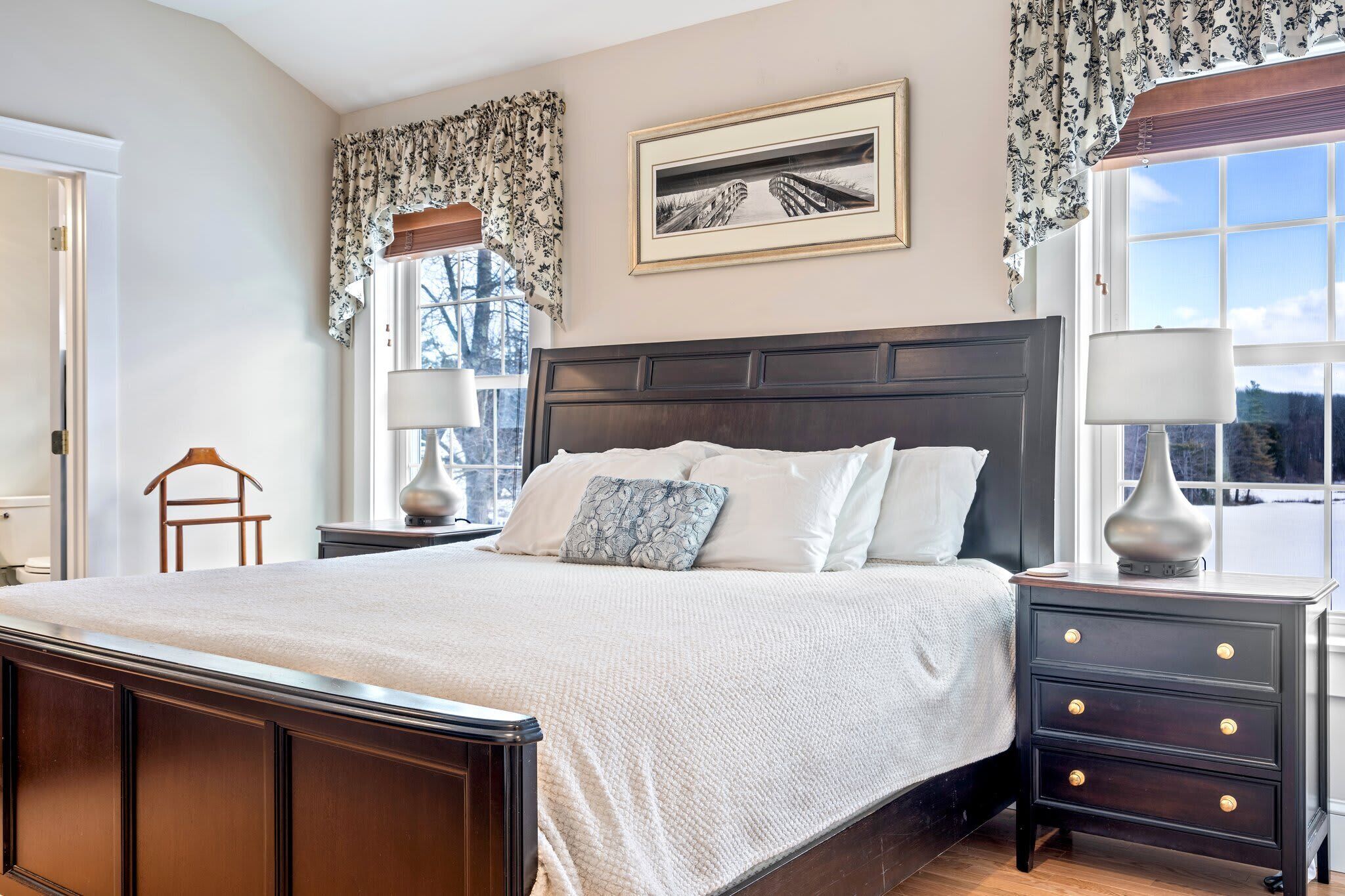 A bedroom with a large bed, dark wooden headboard, white bedding, a nightstand with a lamp, and a window with curtains.