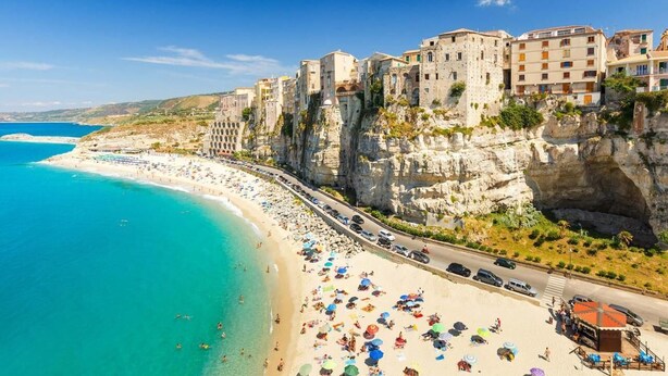 Un pueblo costero con una playa, edificios construidos sobre acantilados y una carretera que bordea la costa.