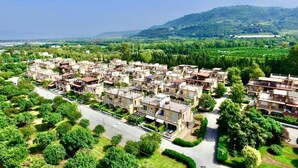 Zona residencial con varias casas de dos pisos, dispuestas en un patrón de cuadrícula, rodeada de vegetación y colinas al fondo.