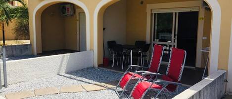 A covered outdoor seating area with red chairs and a table, surrounded by a concrete border and gravel.
