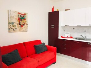Un sofá rojo con dos cojines, una cocina con armarios blancos y una encimera roja, y un cuadro de un búho en la pared.