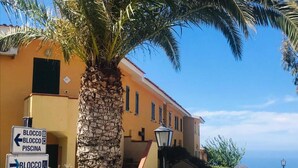 A building with a stone pillar, a palm tree, and directional signs for 'BLOCCO B', 'BLOCCO C', and 'PISCINA'.