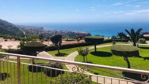 A balcony with a view of a well-maintained garden, a pathway, and a distant coastal town.