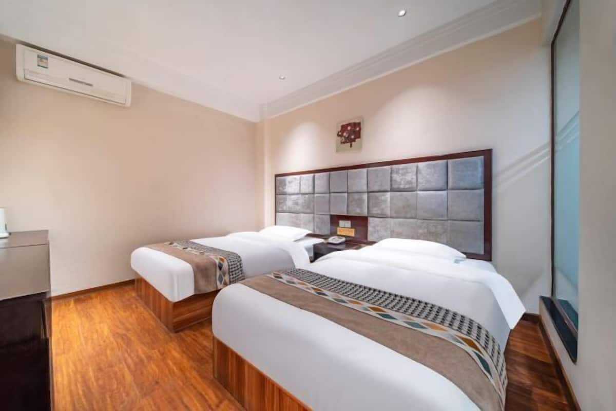 A hotel room with two beds, a wooden floor, a headboard, a small desk, and a wall-mounted air conditioning unit.