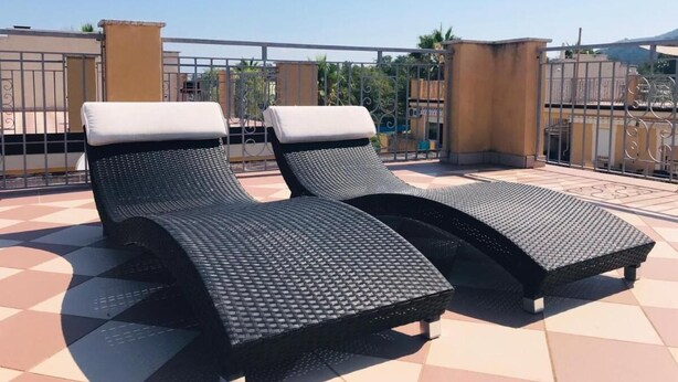 Twee zwarte lounge stoelen met witte kussens op een betegeld terras, met een metalen hek op de achtergrond.