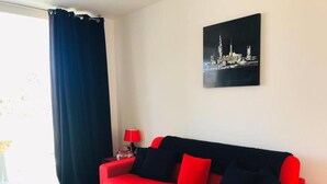 Un sofá rojo con cojines negros en una habitación con pared blanca y un cuadro de un paisaje urbano.