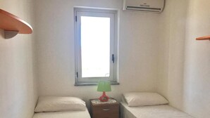 Una habitación pequeña y estrecha con dos camas individuales, una mesita de noche con lámpara y una ventana con persianas.