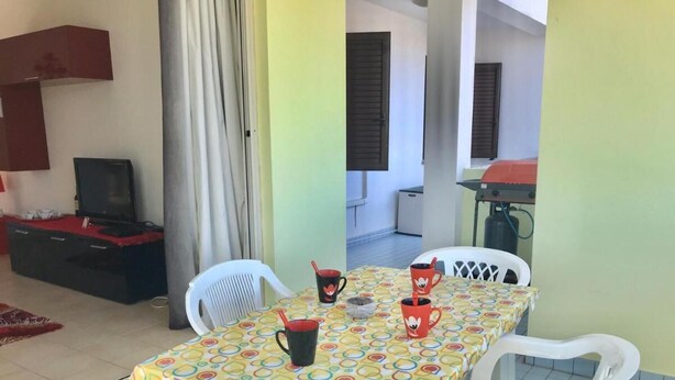 Un comedor con una mesa cubierta por un mantel colorido, sillas de plástico blanco y tazas rojas.