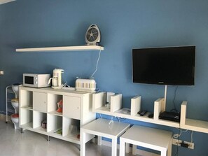 Habitación con una televisión de pantalla plana montada en la pared, un mueble blanco con microondas, cafetera y un ventilador en un estante, y un escritorio blanco con computadora y controles remotos.